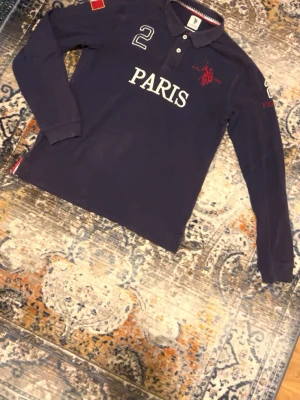 Mörkblå långärmad henley U.S. Polo Assn. - Mörkblå långärmad henley från U.S. Polo Assn. med vita knappar, broderad text 'PARIS' och siffran 2 framtill. Detaljer som franska flaggan på ärmen och röd brodyr med 'FRANCE' och logga. Skön bomullskvalitet och klassisk krage.
