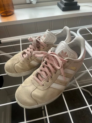 Adidas Gazelle beige med rosa snören - Säljer ett par Adidas Gazelle sneakers i beige mocka med klassiska vita ränder och detaljer. Skorna har rosa snören och vit sula. Adidas-logga i guld på sidan och rosa på hälen. Pris går att pruta ner🤝🏻