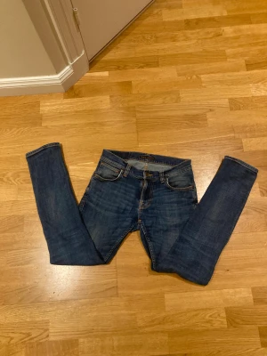 Blå skinny jeans från Nudie Jeans - Snygga blå jeans från Nudie Jeans med klassisk femficksdesign och orangea sömmar. Modellen är skinny med smal passform och raka ben. Jeansen är tillverkade i slitstarkt bomullsmaterial och har en mörk tvätt med lätt slitning på låren.