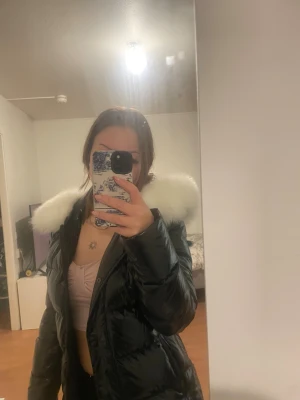 Svart pufferjacka med vit päls - Säljer en svart glansig pufferjacka med fluffig vit äkta päls på huvan. Jackan har quiltad design och dragkedja framtill. Perfekt för dig som vill ha en varm och snygg jacka med lite extra attityd. Passar dig som gillar streetstyle och vill sticka ut.
