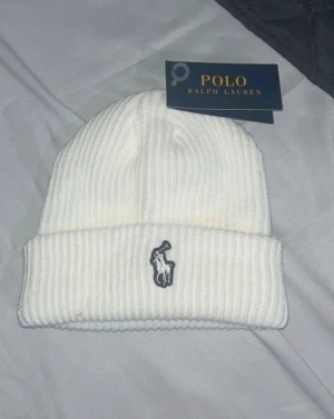 Vit ribbad mössa från Polo Ralph Lauren - Vit ribbstickad mössa från Polo Ralph Lauren med klassisk uppvikt kant och broderad logga framtill. Snygg och enkel design som passar till allt, tillverkad i mjukt stickat material för extra komfort. Pris kan diskuteras 