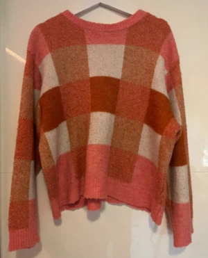 Rutig stickad tröja från Gina Tricot - Supermysig stickad tröja från Gina Tricot med stort rutmönster i rosa, beige och rostbrunt. Tröjan har rund hals, ribbade muddar och lång ärm. Perfekt för dig som gillar färg och vill ha en chill vibe i garderoben.