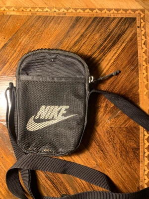 Svart axelväska från Nike - Liten och smidig svart axelväska från Nike med vit logga på framsidan. Väskan har ett meshfack framtill, röd insida och dragkedja upptill. Perfekt för mobilen och småprylar när du är på språng.
