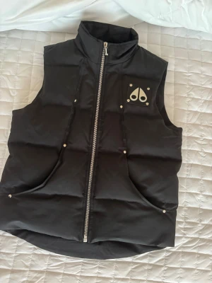 Svart dunväst från Moose Knuckles - Svart dunväst från Moose Knuckles med hög krage och silverfärgad dragkedja framtill. Västen har ett ikoniskt logomärke på bröstet och broderad insida med hockeymotiv. Flera nitar och praktiska fickor. Perfekt för kalla dagar när du vill ha en cool look.