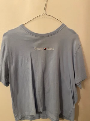 Ljusblå Tommy Jeans t-shirt XL - Snygg ljusblå t-shirt från Tommy Jeans i storlek XL. T-shirten har korta ärmar och är gjord i mjuk bomull. På bröstet finns ett broderat Tommy Jeans-logo med flagga i vitt, rött och blått. Perfekt för en chill och stilren look.