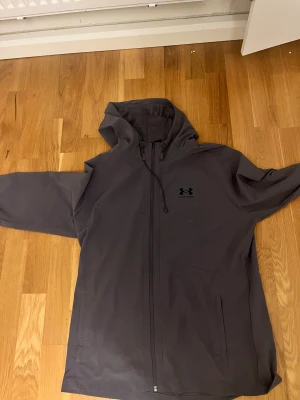 Grå vindjacka från Under Armour - Snygg grå vindjacka från Under Armour med huva och dragkedja hela vägen. Jackan har diskret logga på bröstet och praktiska fickor. Materialet är syntet och jackan har snörning vid huvan. Perfekt för dig som gillar sportig stil och vill ha något funktionellt.