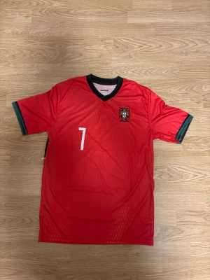 Portugal Ronaldo 7 fotbollströja röd - Snygg röd Portugal fotbollströja med nummer 7 och Ronaldo på ryggen. Tröjan har gröna detaljer vid ärmslut och svart krage. Tillverkad i lätt och ventilerande material, perfekt för match eller träning. Klassisk passform och officiellt klubbmärke på bröstet.