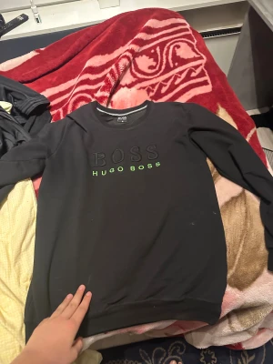 Svart Hugo Boss sweatshirt - Svart sweatshirt från Hugo Boss med broderad BOSS-text och neongrön HUGO BOSS-logga på bröstet. Tröjan har rund hals, långa ärmar och ribbade muddar vid ärmslut och nederkant. Perfekt för dig som gillar stilrena märkesplagg.