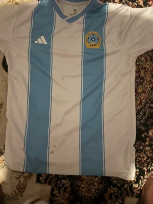 Somalia SFF fotbollströja Adidas  - Somalia Football Federation matchtröja från Adidas i storlek XXL. Tröjan är vit med ljusblå vertikala ränder, broderat SFF-märke på bröstet och klassisk Adidas-logga. Tillverkad i lätt och ventilerande polyester, perfekt för fotbollsträning eller samling.