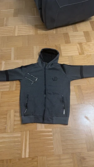 Grå hoodie från Philipp Plein L - Snygg mörkgrå hoodie från Philipp Plein i storlek L. Tröjan har huva, hel dragkedja framtill och coola detaljer med extra dragkedjor på bröstet. Märkets logga sitter på vänster sida. Perfekt för dig som gillar streetwear och vill sticka ut.