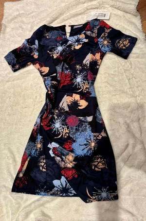 Blommig kortklänning från Fransa - Supersnygg kortklänning från Fransa med färgstarkt blommönster i rött, blått, beige och svart. Klänningen har korta ärmar, rund halsringning och är gjord i ett mjukt, följsamt polyestertyg. Perfekt för dig som vill sticka ut med en cool och modern look. Kappan och väskan som syns i bild är också till salu på samma adress. (Miss sixty & Karen millen)