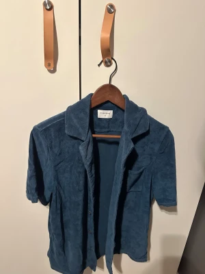 Frottéskjorta  - Mjuk och chill blå skjorta från Studio Total i frotté. Skjortan har korta ärmar, klassisk krage, bröstficka och knappar framtill. Perfekt för dig som gillar en avslappnad vibe och vill sticka ut med något annorlunda.