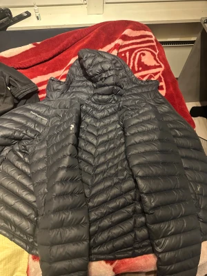 Svart dunjacka med huva från Peak Performance - Säljer en svart dunjacka från Peak Performance med snyggt quiltad design och huva. Jackan har dragkedja framtill och är tillverkad i lätt och slitstark polyester. Perfekt för kalla dagar och har en clean look som funkar till det mesta. Den har också några hål.