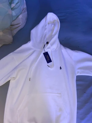 Vit hoodie från Polo Ralph Lauren - Vit hoodie från Polo Ralph Lauren med klassisk broderad logga på bröstet. Tröjan har huva med snörning, ribbade muddar och en stor magficka. Materialet är mjuk bomull och passformen är normal. Perfekt för chill dagar och enkel att matcha med jeans eller joggers.