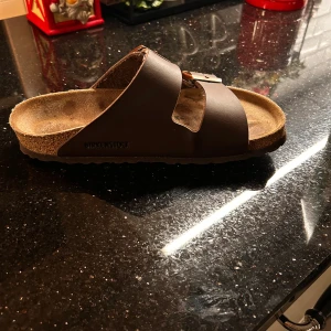 Bruna Birkenstock sandaler med spännen - Säljer ett par klassiska Birkenstock sandaler i mörkbrunt skinn med två breda remmar och metallspännen. Sulan är ergonomiskt formad med kork och gummibotten för extra komfort. Perfekt för chill dagar och enkel stil.