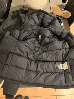 Svart dunjacka Canada Goose med päls - Säljer en svart dunjacka från Canada Goose med fluffig beige päls på huvan. Jackan har broderad logga på ärmen, dragkedja och knappar framtill samt stora fickor. Perfekt för kalla vinterdagar och har en riktigt schysst oversized look.