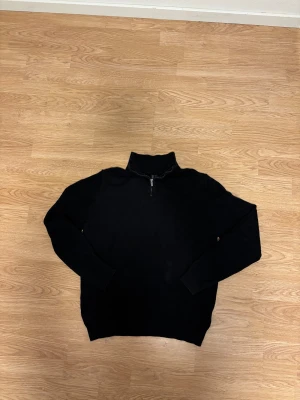 Svart stickad tröja med half zip - Svart stickad tröja med halv dragkedja i halsen. Tröjan har långärmade ärmar och en enkel, clean design som funkar till många olika stilar. Perfekt för dig som gillar basic men ändå vill ha något med lite edge.