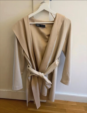 Beige trenchcoat med bälte från Zara - Snygg beige trenchcoat från Zara med bred krage och knytbälte i midjan. Jackan har långa ärmar och två stora fickor framtill. Perfekt för dig som gillar stilrena och minimalistiska plagg med en modern touch. Nästan Aldrig använd!!!