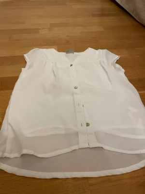 Vit kortärmad skjorta från Name It - Fräsch vit skjorta från Name It med korta ärmar och knappar framtill. Skjortan har en lätt och luftig känsla, rundad nederkant och diskreta detaljer vid axlarna. Perfekt för dig som gillar enkel och stilren look.