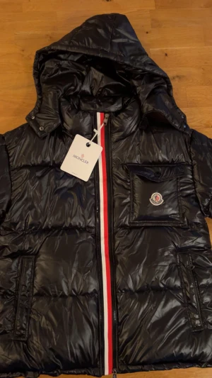 Svart Moncler pufferjacka med huva - Svart pufferjacka från Moncler med glansig finish, huva och dragkedja framtill med röd och vit rand. Jackan har en bröstficka med Moncler-logga, två sidofickor med dragkedja och quiltad design. Perfekt för kalla dagar och riktigt snygg streetstil.