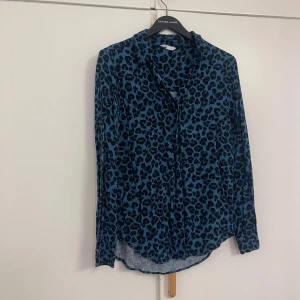 Blå leopardmönstrad skjorta S - Blå skjorta med svart leopardmönster från C i storlek S. Skjortan har klassisk krage, knappar framtill och långa ärmar med knappslå vid manschetten. Materialet är mjukt och lätt, perfekt för dig som vill sticka ut med ett coolt mönster.