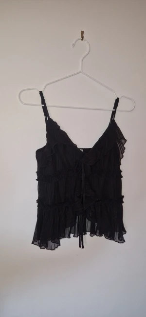 Svart blus  - Säljer en svart blus från Amisu i storlek M. Toppen har tunna axelband, volanger och plisserade detaljer samt en söt knytning framtill. Materialet är lätt och transparent  som ger en luftig känsla. Perfekt för dig som gillar romantiska och trendiga plagg. 