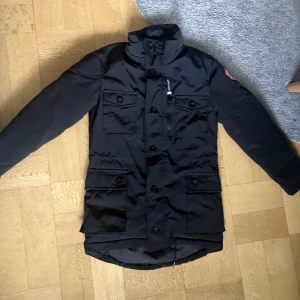 Svart parkajacka från Canada Goose - Svart parkajacka från Canada Goose med hög krage, flera fickor med knappar och dragkedja på bröstet. Klassisk röd och vit Canada Goose-patch på ärmen. Jackan har lång ärm och en stilren, funktionell design som passar perfekt när det är kallt ute. Storlek 8 i barn storlek vilket motsvara ungefär XS i vuxen storlek