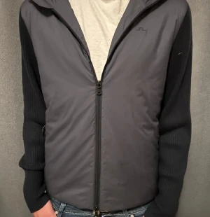 Svart hybridjacka från J.Lindeberg - Svart jacka från J.Lindeberg med stickade ärmar och rygg i ull, samt quiltad framsida och huva i syntetmaterial. Jackan har hel dragkedja framtill och en stilren, sportig look. Perfekt för dig som gillar streetwear med en twist.                                      Storlek S men passar även M.                                           Ny priss 3000 kr mitt priss 1000 kr!