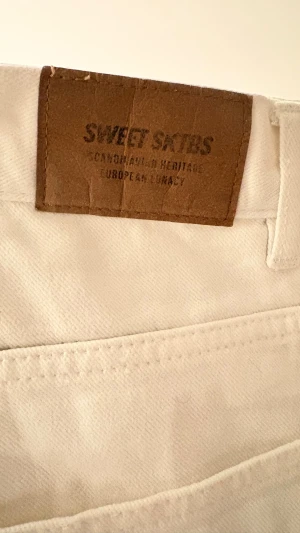 Vita loose shorts från Sweet Skies - Säljer ett par vita shorts från Sweet Skies med loose passform och coolt Peace-märke broderat på benet. De har praktiska fickor och är gjorda i mjuk bomull, perfekta för chill dagar. Midjan är midwaist och tyget är slätt och bekvämt.