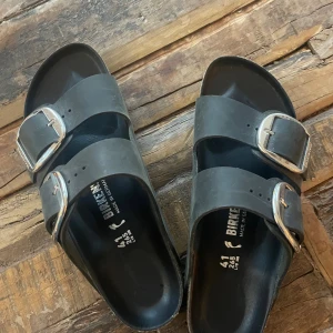 Svarta Birkenstock sandaler med spännen - Birkenstock sandaler i svart med två breda remmar och stora silvriga spännen. Sulan är ergonomisk med korkdetaljer och mönstrad undersida för extra grepp. Remmarna är i läder och spännena har Birkenstock-logga. Perfekt för chill dagar under sommaren.
