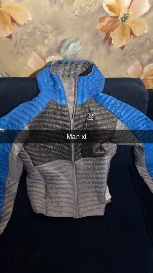 Grå och blå pufferjacka Haglöfs XL man - Säljer en grå och blå pufferjacka från Haglöfs i storlek XL för herr. Jackan har huva, hel dragkedja och quiltad design som ger en sportig vibe. Perfekt för kyliga dagar och outdoor-aktiviteter. Skön passform och lätt att bära. Med prislapp