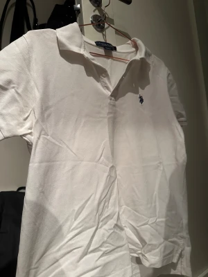 Vit pikétröja från U.S. Polo Assn - Säljer en klassisk vit pikétröja från U.S. Polo Assn med korta ärmar och krage. Tröjan har en liten broderad logga på bröstet och är tillverkad i 100% bomull, vilket gör den skön och luftig. Perfekt för dig som gillar stilrena och sportiga plagg.