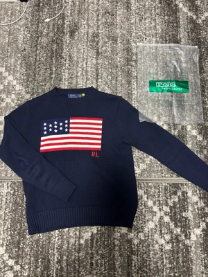 Mörkblå stickad tröja Polo Ralph Lauren - Snygg mörkblå stickad tröja från Polo Ralph Lauren med amerikansk flagga i rött, vitt och blått på bröstet samt RL-broderi. Rund hals och ribbade muddar vid ärmslut och nederkant. Perfekt för dig som gillar klassisk stil med en twist. Använts 2-3 gånger