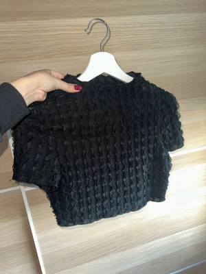 Partytopp med struktur - Säljer en svart croppad party topp med korta ärmar och strukturerat tyg. Oanvänd och i topp skick. Saknar storleksangivelse men motsvarar ungefär S! 