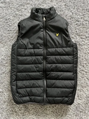 Svart dunväst från Lyle & Scott Junior - Snygg svart dunväst från Lyle & Scott Junior med gul broderad logga på bröstet. Västen har quiltad design, hög krage och dragkedja framtill. Perfekt att slänga över en hoodie när det är kyligt ute. Passar dig som gillar stilren och sportig look.