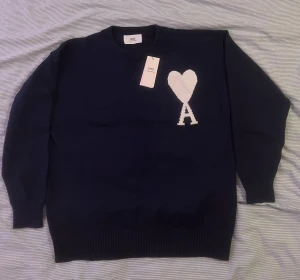 Mörkblå stickad tröja från Ami - Snygg mörkblå stickad tröja från Ami med ett stort vitt hjärta och bokstaven A på bröstet. Tröjan har rund hals, ribbade muddar och lång ärm. Perfekt för dig som gillar stilrena detaljer och vill ha något som sticker ut. Perfekt för vintern