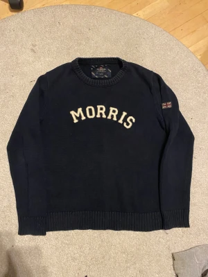 Mörkblå stickad tröja Morris S - Snygg mörkblå stickad tröja från Morris i storlek Small. Tröjan har vit broderad MORRIS-text på bröstet och en patch med brittiska flaggan på ärmen. Rund hals och ribbade muddar vid ärmslut och nederkant. Perfekt för dig som gillar klassisk stil.