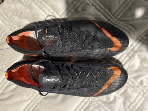 Nike Mercurial Vapor360 fotbollsskor - Säljer ett par svarta Nike Mercurial Vapor360 fotbollsskor med orange detaljer och snörning. Skorna har en låg profil, rund tå och platt sula med metall-dobbar för grymt grepp på planen. Ovandelen är i syntetmaterial och insidan är orange. Perfekt för dig som vill ha speed och kontroll.