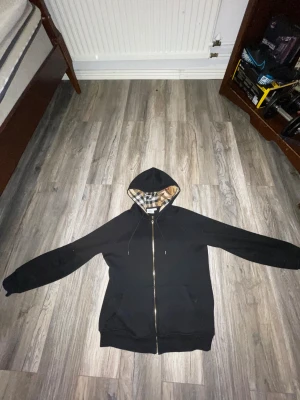 Svart Burberry kofta  - Svart hoodie från Burberry i storlek XS/S  med klassisk rutig insida på huvan i beige, svart och vit. Tröjan har dragkedja i guld, snörning med metalländar och två fickor framtill. Materialet är mjuk bomull och polyester, perfekt för chill dagar.