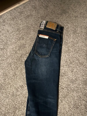 Mörkblå Nudie Jeans, storlek 28/30 - Snygga mörkblå jeans från Nudie Jeans med klassisk femficksdesign och orangea sömmar. Modellen har en rak passform och är tillverkad i slitstarkt denimtyg. Perfekt för dig som gillar stilrena och tidlösa jeans med coola detaljer.