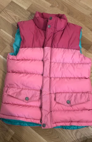 Rosa och röd dunväst från Everest - Snygg dunväst från Everest i färgblockad design med mörkröd upptill och ljusrosa nedtill. Västen har två fickor med tryckknappar, hög krage och turkos insida. Materialet är 100% polyester och den stängs med dragkedja och tryckknappar framtill.