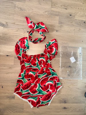 Röd body med vattenmelonmönster Vattenmelon Romper med Hårband - Superfin body med färgglatt vattenmelonmönster i rött, grönt och svart. Modellen har puffiga axelband, elastisk midja och vit spetskant nertill. Matchande hårband ingår för en extra söt look. Perfekt för speciella tillfällen! 🍉 Ett sött set med en romper och ett matchande hårband med vattenmelonsmönster. Romperen har volangärmar och en spetskant. Storlek 9-18 mån