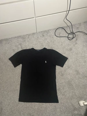 Svart t-shirt från Polo Ralph Lauren - Svart t-shirt från Polo Ralph Lauren med klassisk passform och vit broderad logga på bröstet. Gjord i mjuk bomull och har rund halsringning samt korta ärmar. Perfekt basic-plagg med stilren look. Pris kan diskuteras!