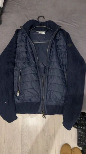 J.Lindeberg Cardigan - Snygg mörkblå cardigan från J.Lindeberg i storlek M. Cardiganen har quiltad front och stickade ärmar och sidor. Två dragkedjeförsedda fickor och grov ribbad krage ger en cool look. Perfekt för höst och vinter, med stilren design och diskret logopatch på ärmen. Storlek M. PRISET HUGGS EJ I HALV.