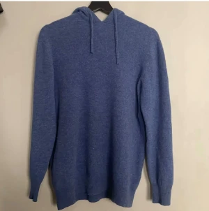 Blå stickad hoodie med huva - Mysig blå hoodie med stickad känsla och huva med snörning. Tröjan har långa ärmar och ribbade muddar vid ärmslut och nederkant. Perfekt för dig som gillar chill och enkel stil. Färgen är en snygg mellanmörk blå som funkar året runt. Liten i storlek, så har du S funkar denna också 