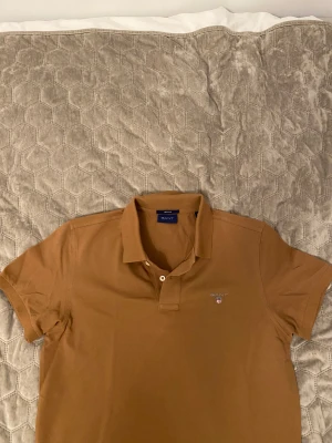 Brun pikétröja från GANT - Snygg brun pikétröja från GANT med klassisk krage och två knappar framtill. Tröjan har korta ärmar och GANT-logga broderad på bröstet. Perfekt för dig som gillar stilrena och tidlösa plagg. Materialet är mjuk bomull som känns skönt mot huden.