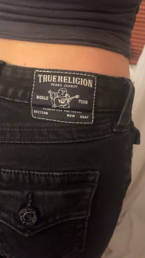 True Religion svarta bootcut jeans model Becca - Svarta jeans från True Religion, modell Becca Mid Rise Bootcut. Snygg bootcut-passform med medelhög midja och klassiska fickor med knappdetalj bak. Jeansen har en cool logopatch och är tillverkade i stretchigt denim för skön passform. Köpta i vintras, knappt använda, fint skick.