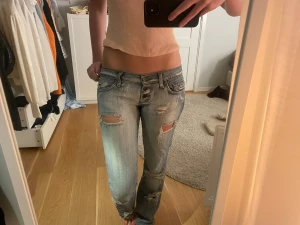 Blå ripped jeans från Sexy Woman - Säljer ett par ljusblå jeans från Sexy Woman med slitna detaljer och flera revor både fram och bak. Modellen är lågmidjad med raka ben och klassisk femficksdesign. Jeansen har knappar i midjan och är tillverkade i jeansmaterial. Jeansen är lite stora i storleken. Midjemåttet är 84cm.