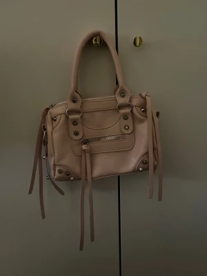 Beige handväska med dragkedja - Snygg beige handväska i skinnimitation med silverfärgade detaljer och dragkedja framtill. Väskan har två handtag, dekorativa nitar och långa remmar som ger en cool vibe. Perfekt storlek för det viktigaste och passar till många olika outfits.