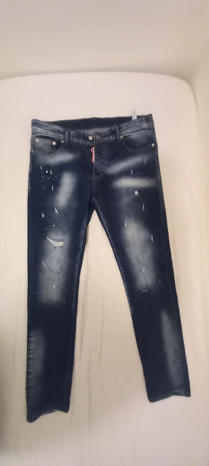 Dsquared2 blå jeans med slitningar - Snygga blå jeans från Dsquared2 i modell Cool Guy med slitna detaljer och färgstänk. Jeansen har klassisk femficksdesign, knappgylf och Dsquared2-logga på röd etikett. Materialet är bomull och passformen är smal med avsmalnande ben.